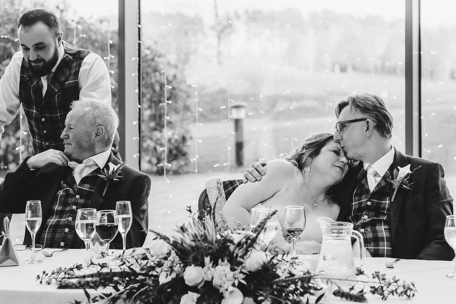 Black and white wedding photo of couples seated at reception table with floral centerpiece.