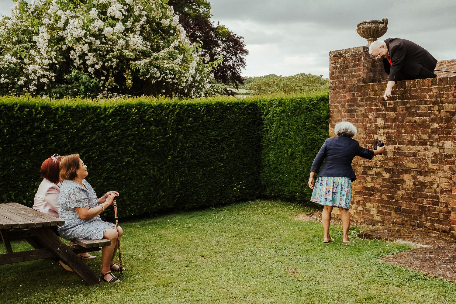 People enjoying a casual outdoor gathering near a brick wall and garden hedge.