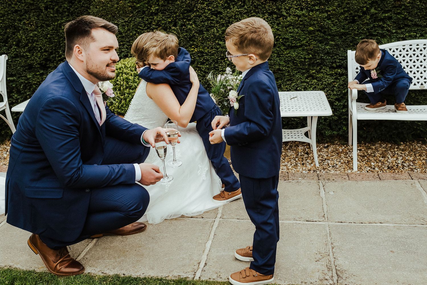 Groom in navy suit kneels to interact with young boys at outdoor wedding.
