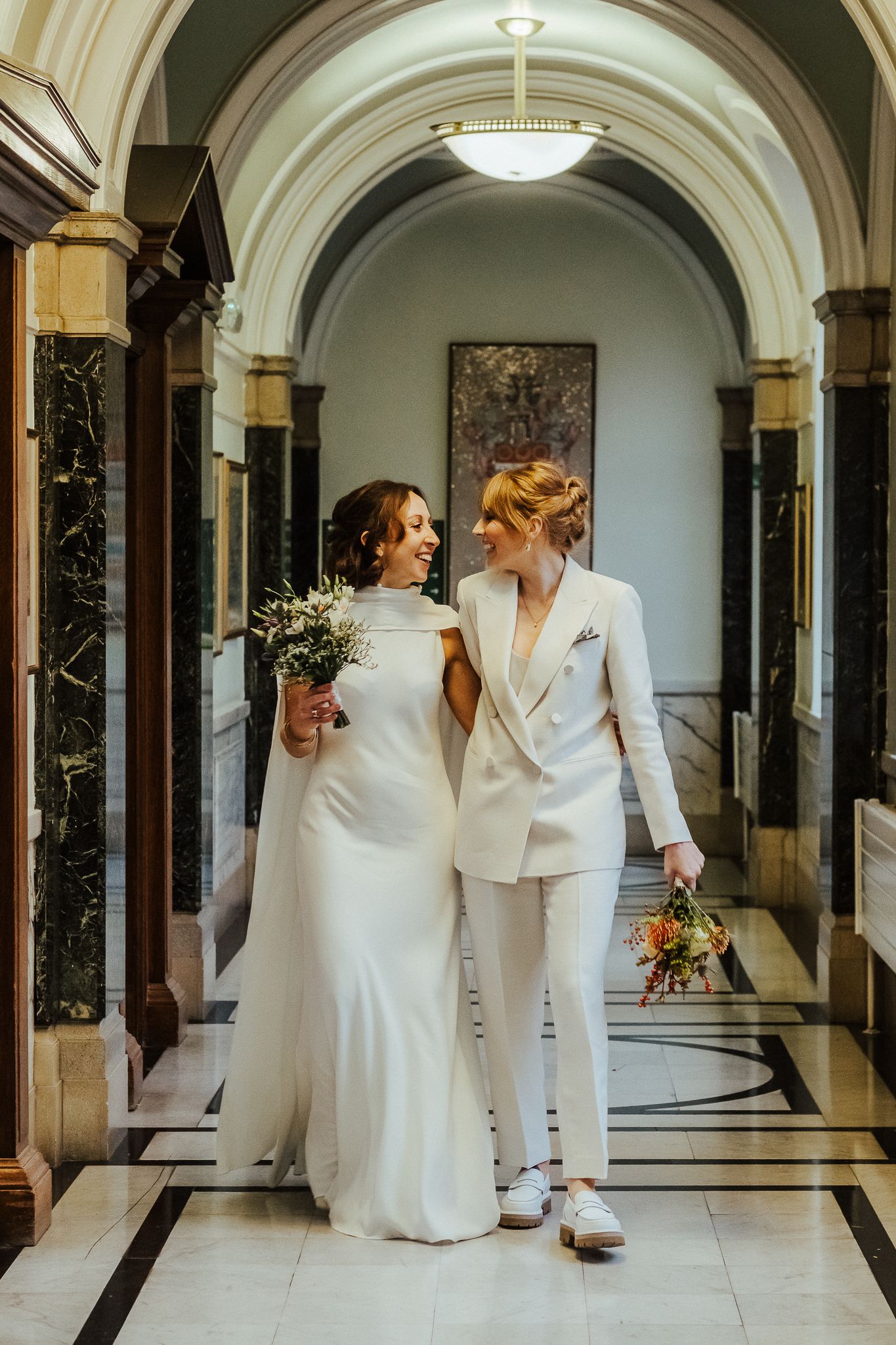 Two people in wedding attire walk together in an elegant hallway with marble columns and arched ceilings.