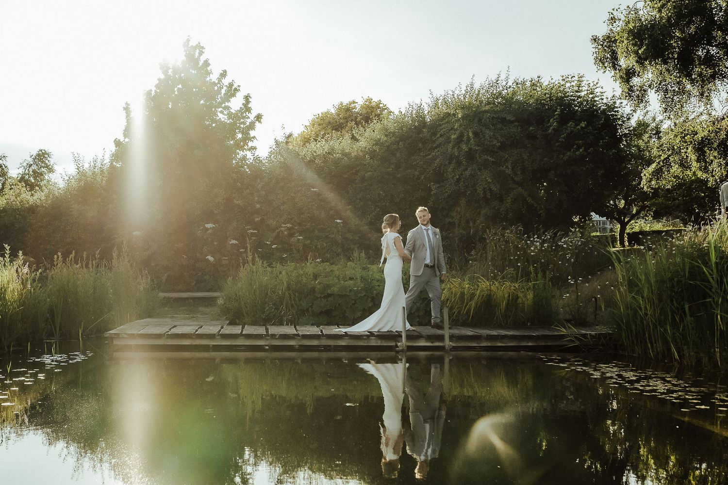 A couple standing together on a wooden dock beside a tranquil pond at sunset, creating a romantic reflection.