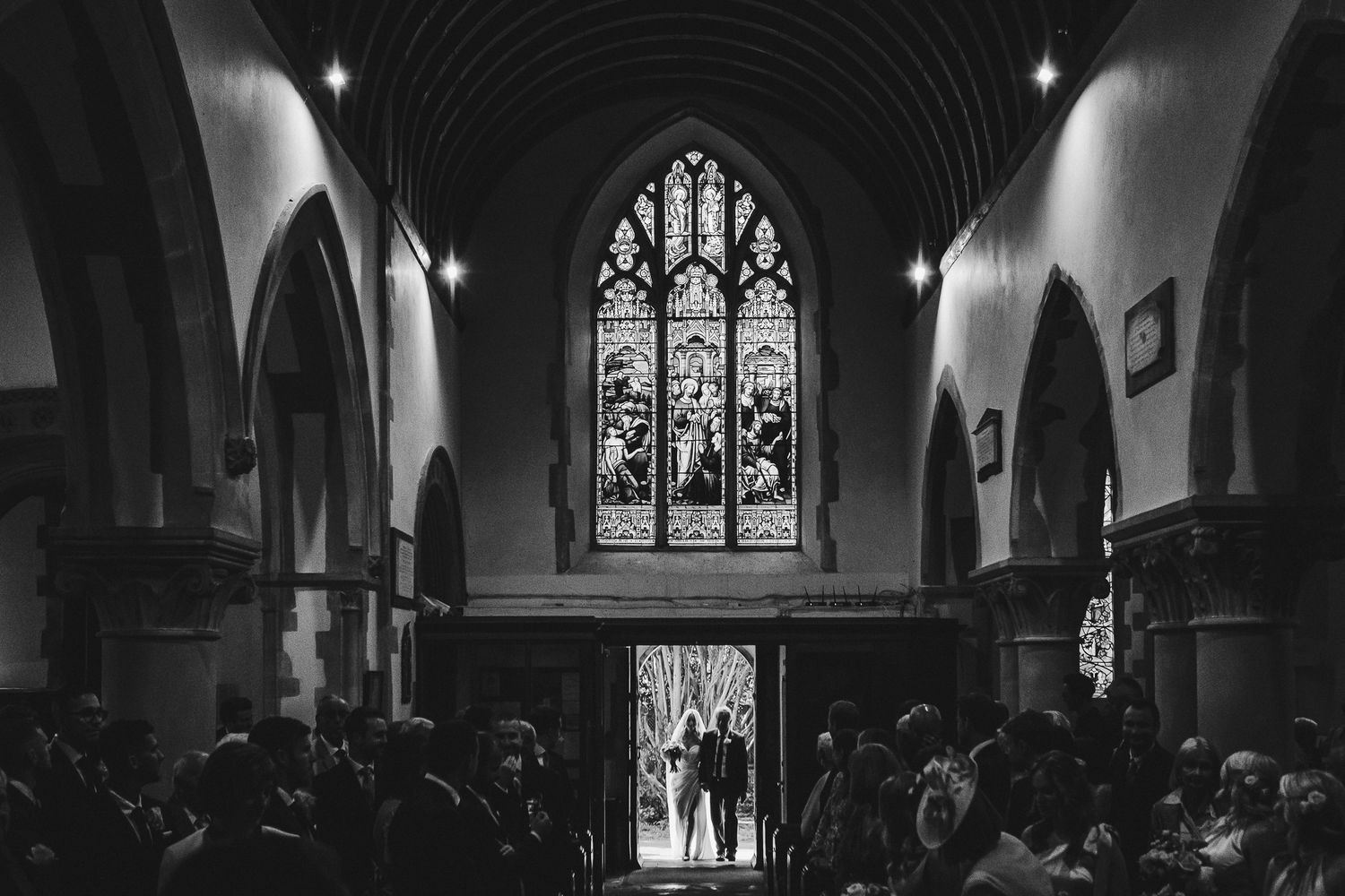 A black and white photo of wedding guests in a church with stained glass windows.
