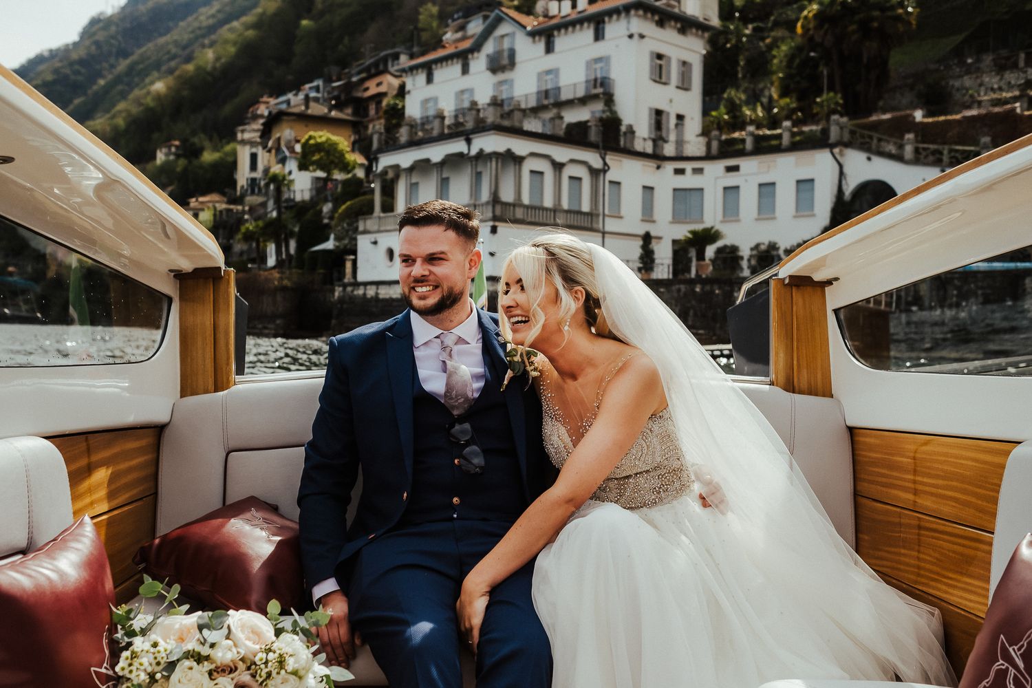 Couple relaxing in wooden boat with Lake Como's picturesque shoreline in background.