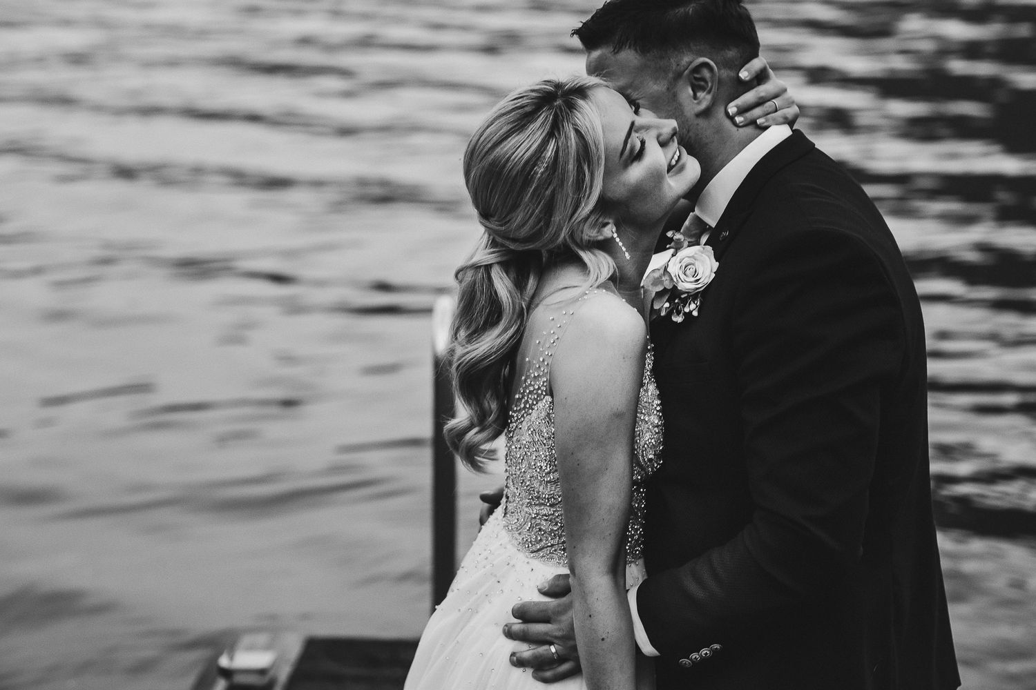 Black and white intimate wedding photo of a couple embracing on a lakeside dock.