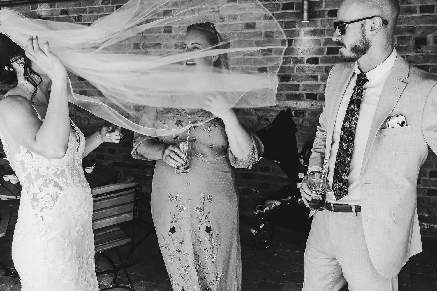 Wedding party playfully posing with flowing veil against brick wall background.