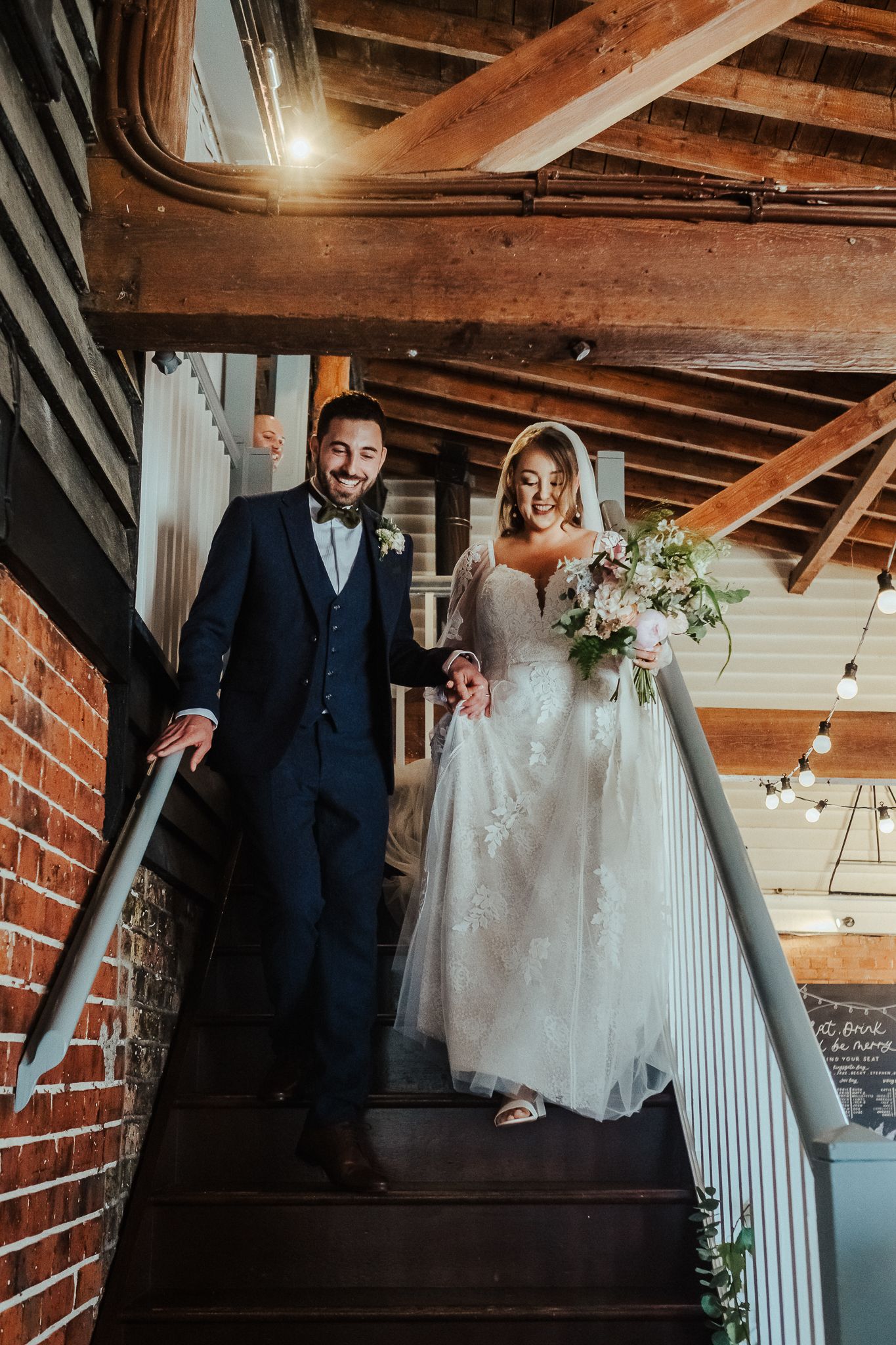Newlyweds pose on rustic staircase with exposed brick walls and wooden beams.