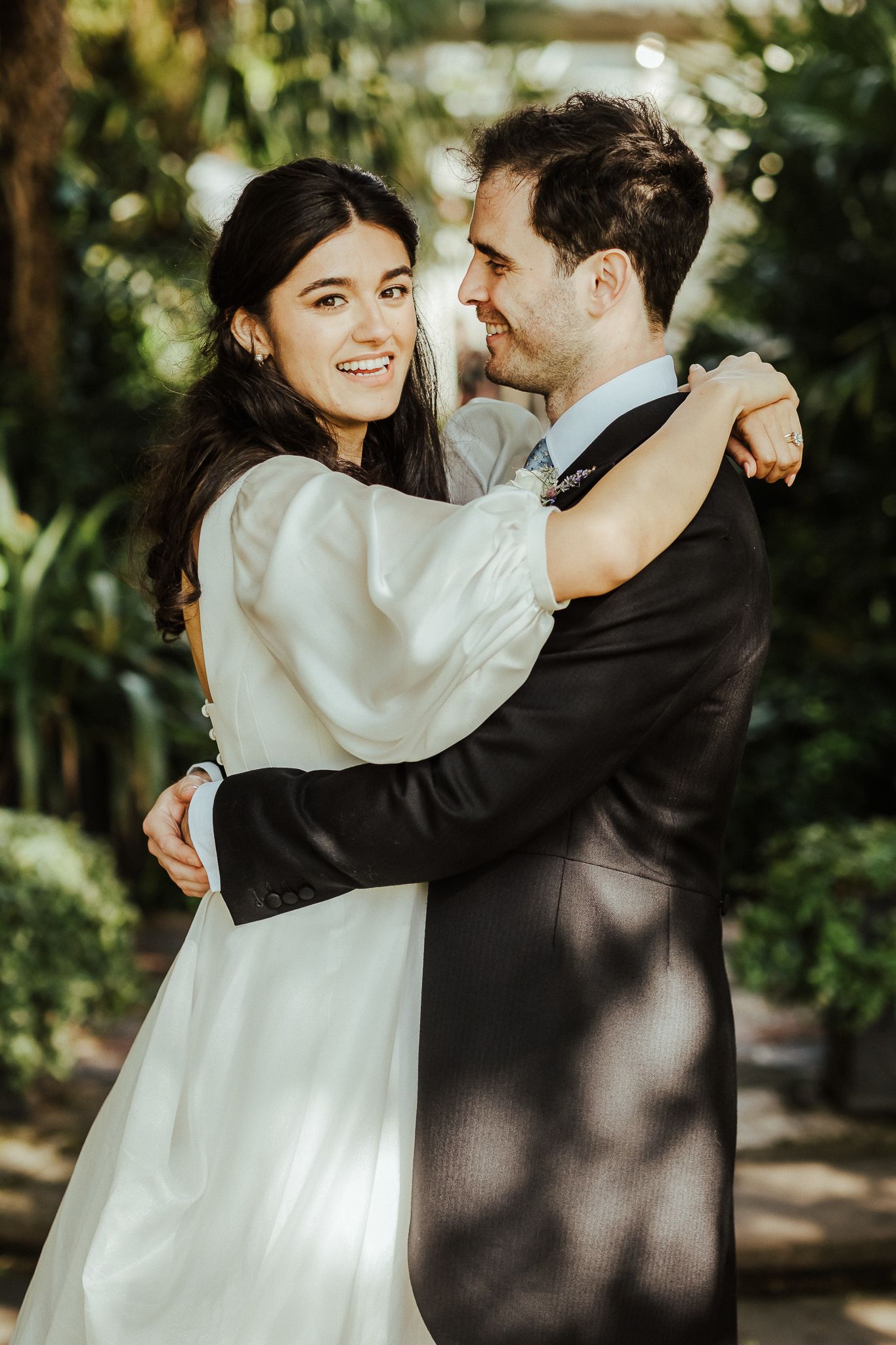 Joyful bride and groom embracing outdoors in dappled sunlight filtering through trees.