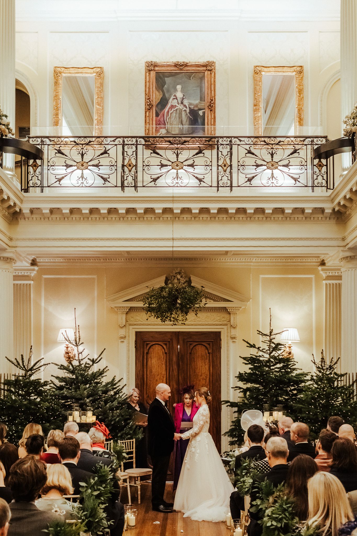 Grand wedding ceremony in historic hall with Christmas trees and ornate balcony.