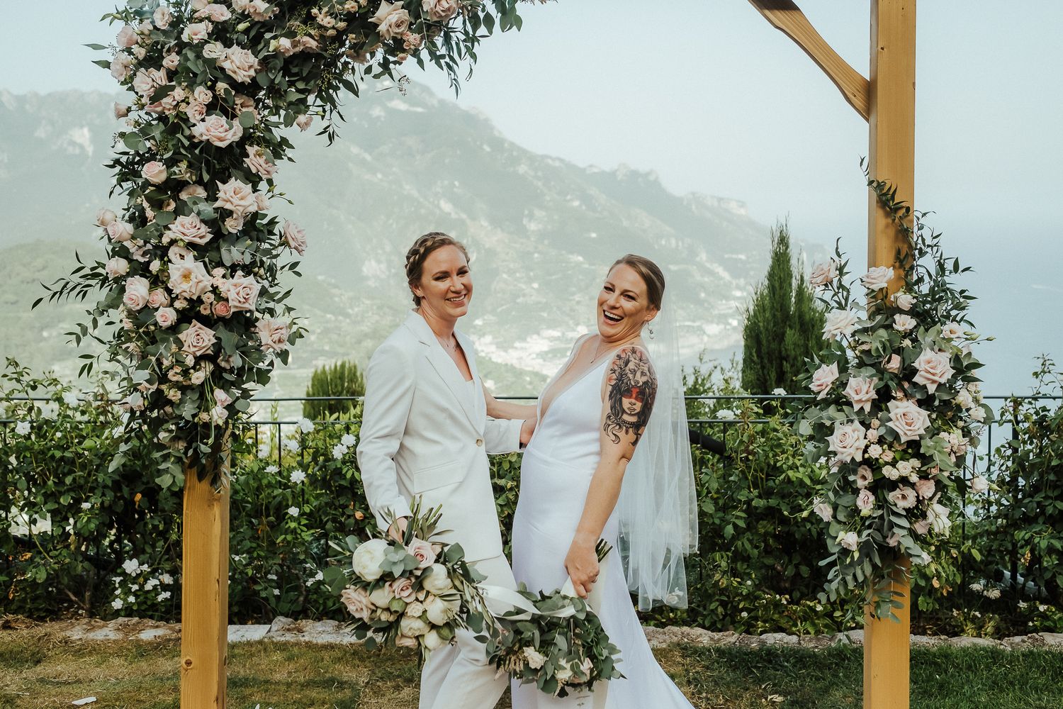 A couple shares a joyful moment under a floral arch with mountain views during their outdoor wedding ceremony.