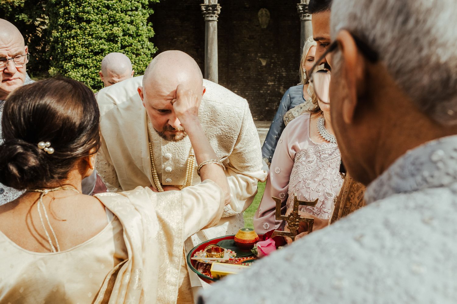A group of people participate in a traditional wedding ceremony ritual outdoors next to a decorative stone pillar.