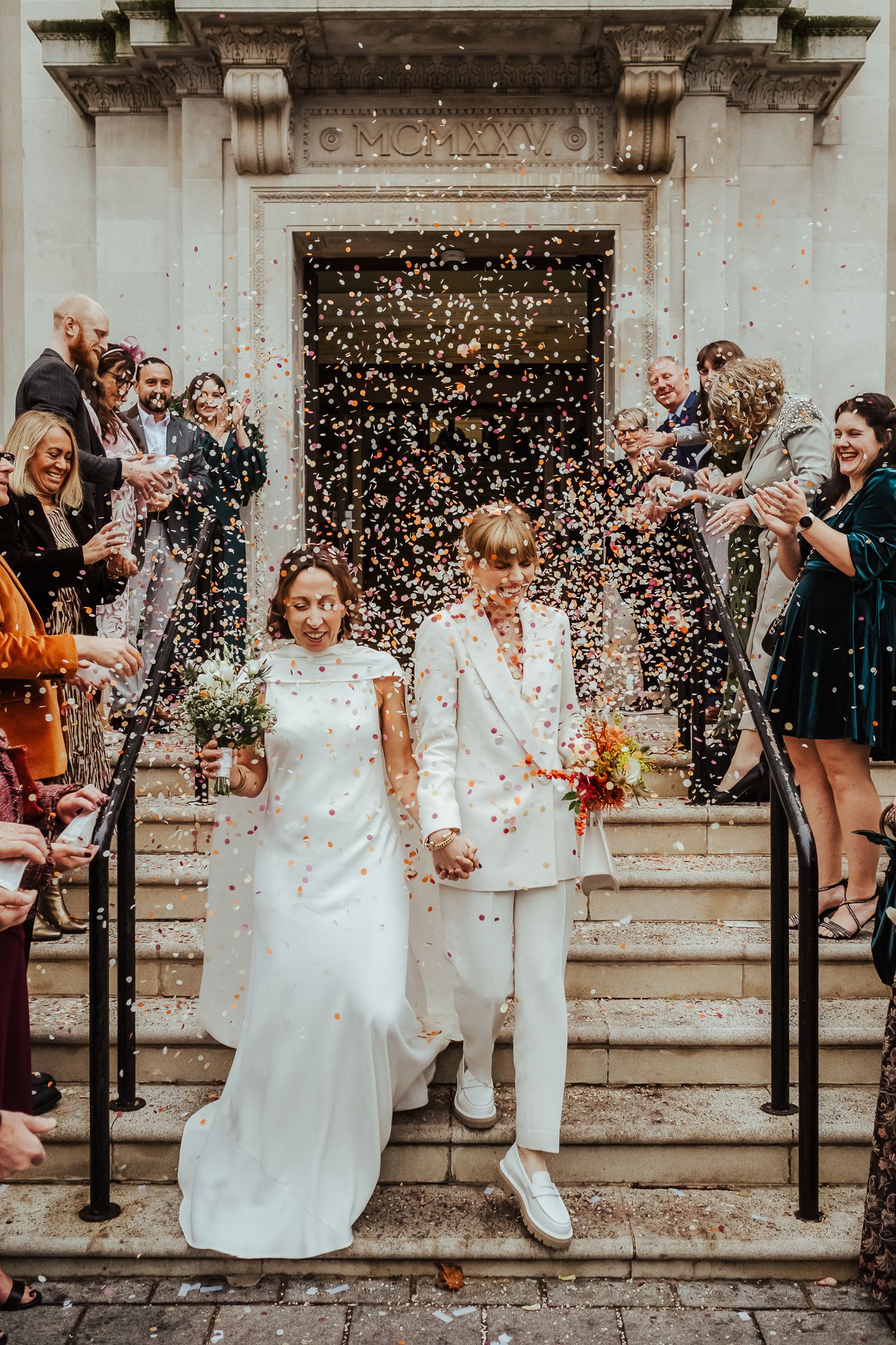 Wedding couple exits venue as guests shower them with confetti on stone steps.