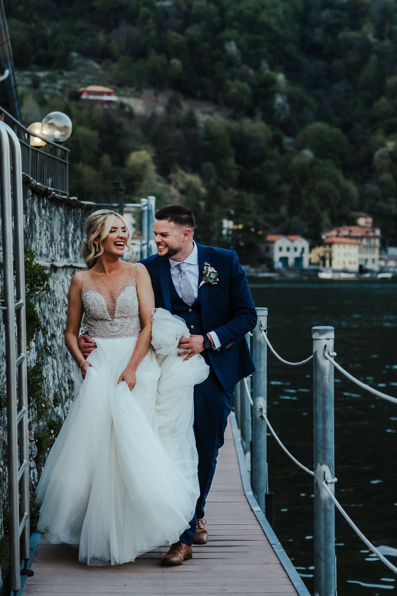 Joyful couple walking along a lakeside dock in elegant wedding attire.