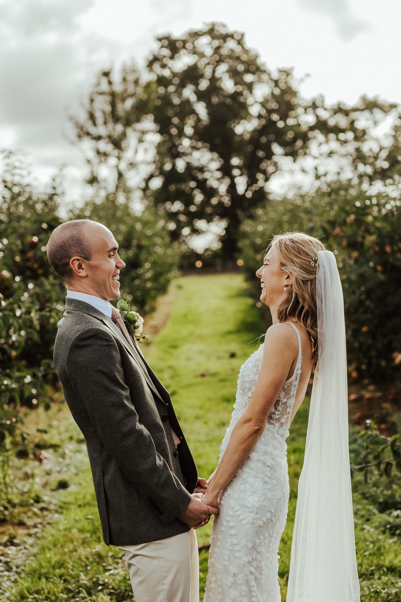 Bride and groom share a laughing moment in orchard during wedding portraits.