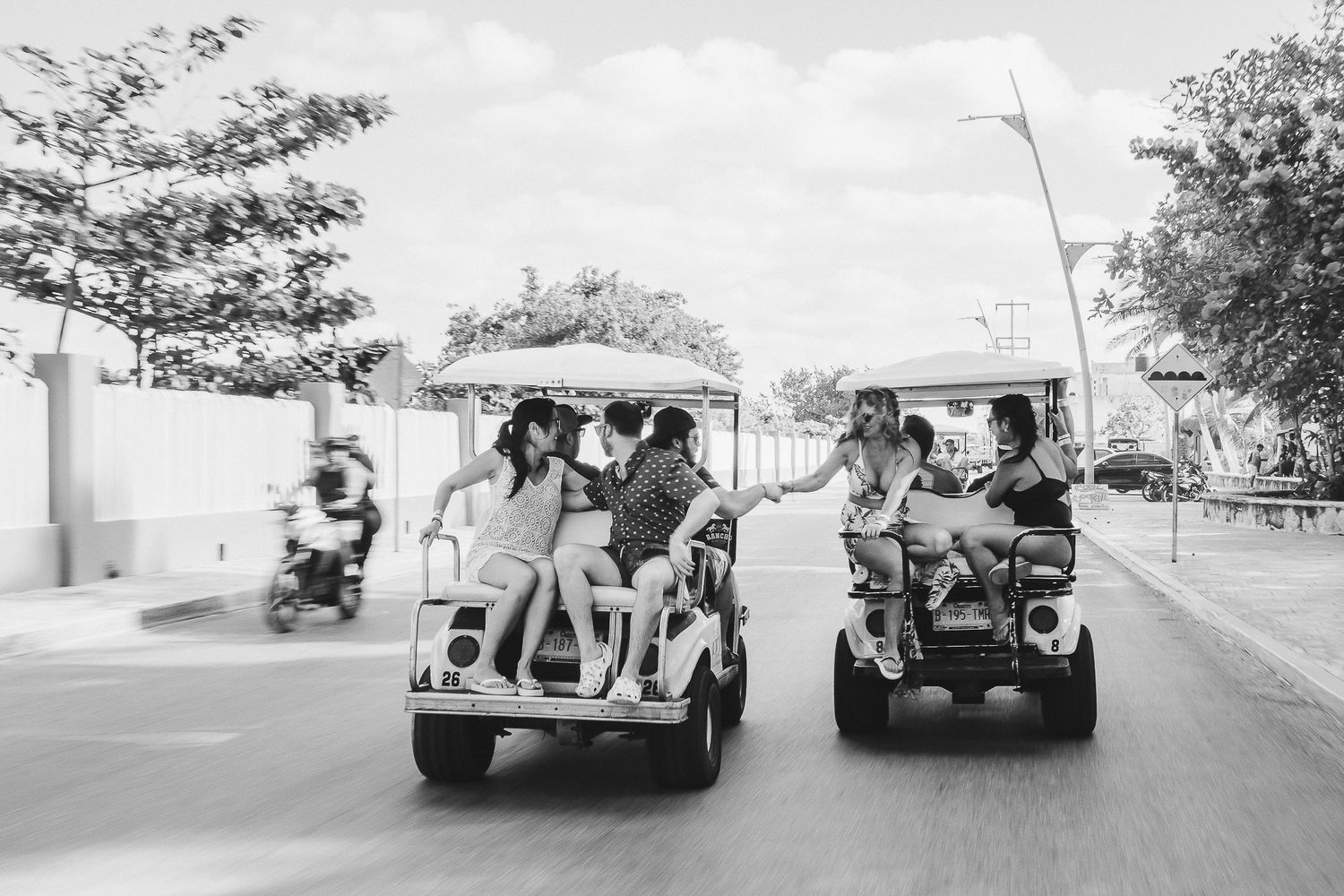 Groups riding in golf carts on a sunny street in black and white.