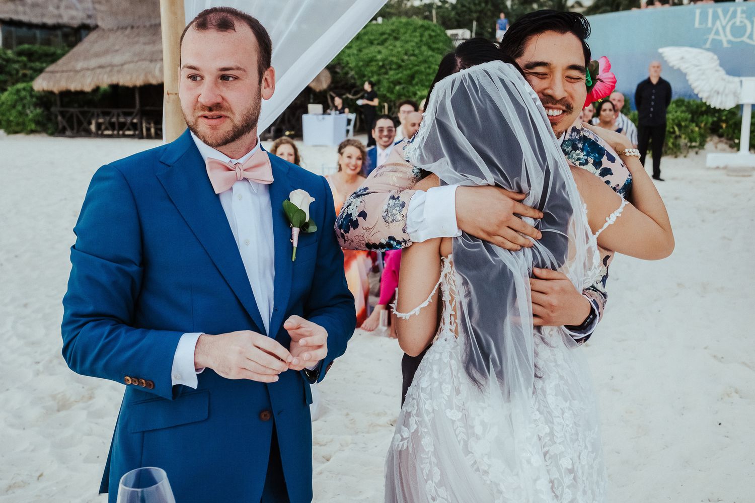 Beach wedding celebration with guests in formal attire under a white canopy.