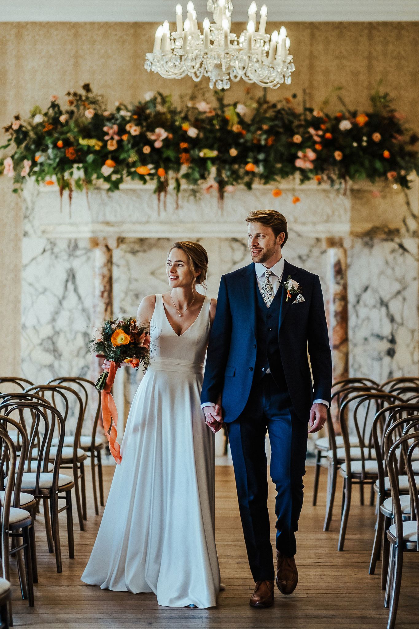 Elegant ceremony space with floral arch and chandelier as couple walks through.