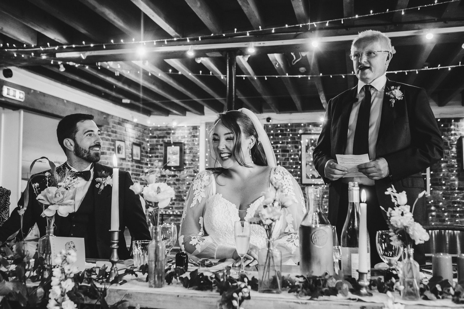 Black and white reception scene with guests giving speeches at decorated wedding table.