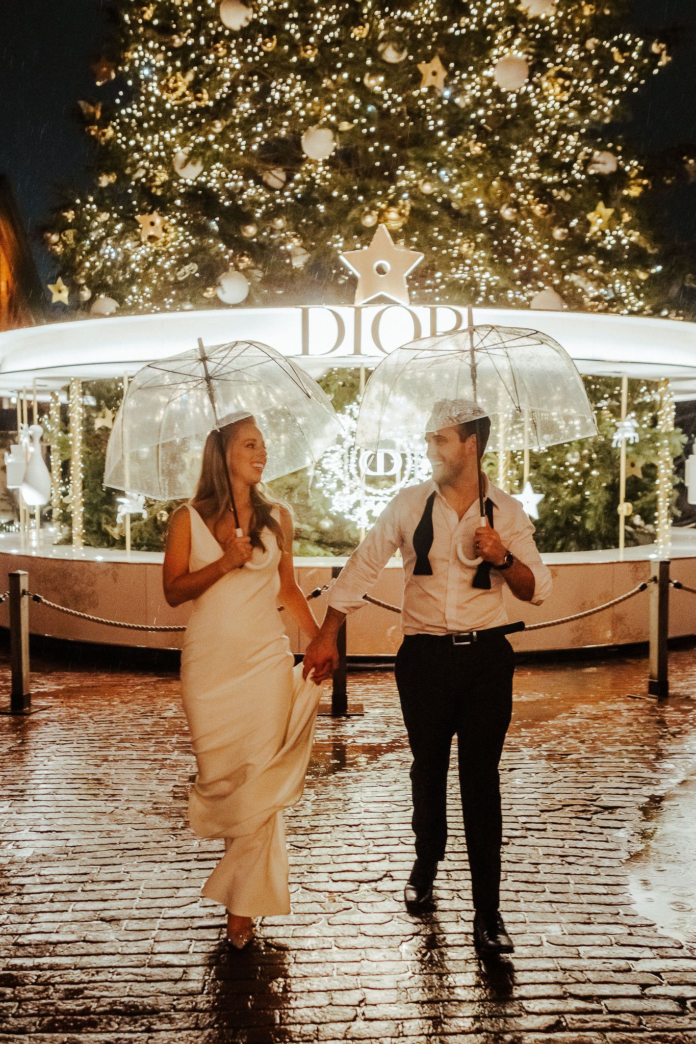 Couple laughs under umbrella by illuminated Dior holiday display.