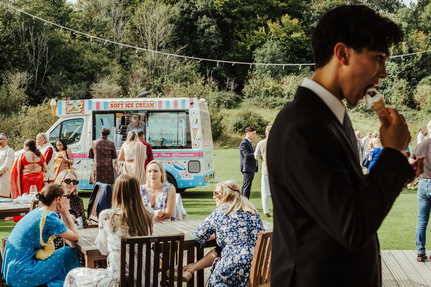 Wedding guests enjoy outdoor reception with ice cream truck and garden party atmosphere on sunny summer day.
