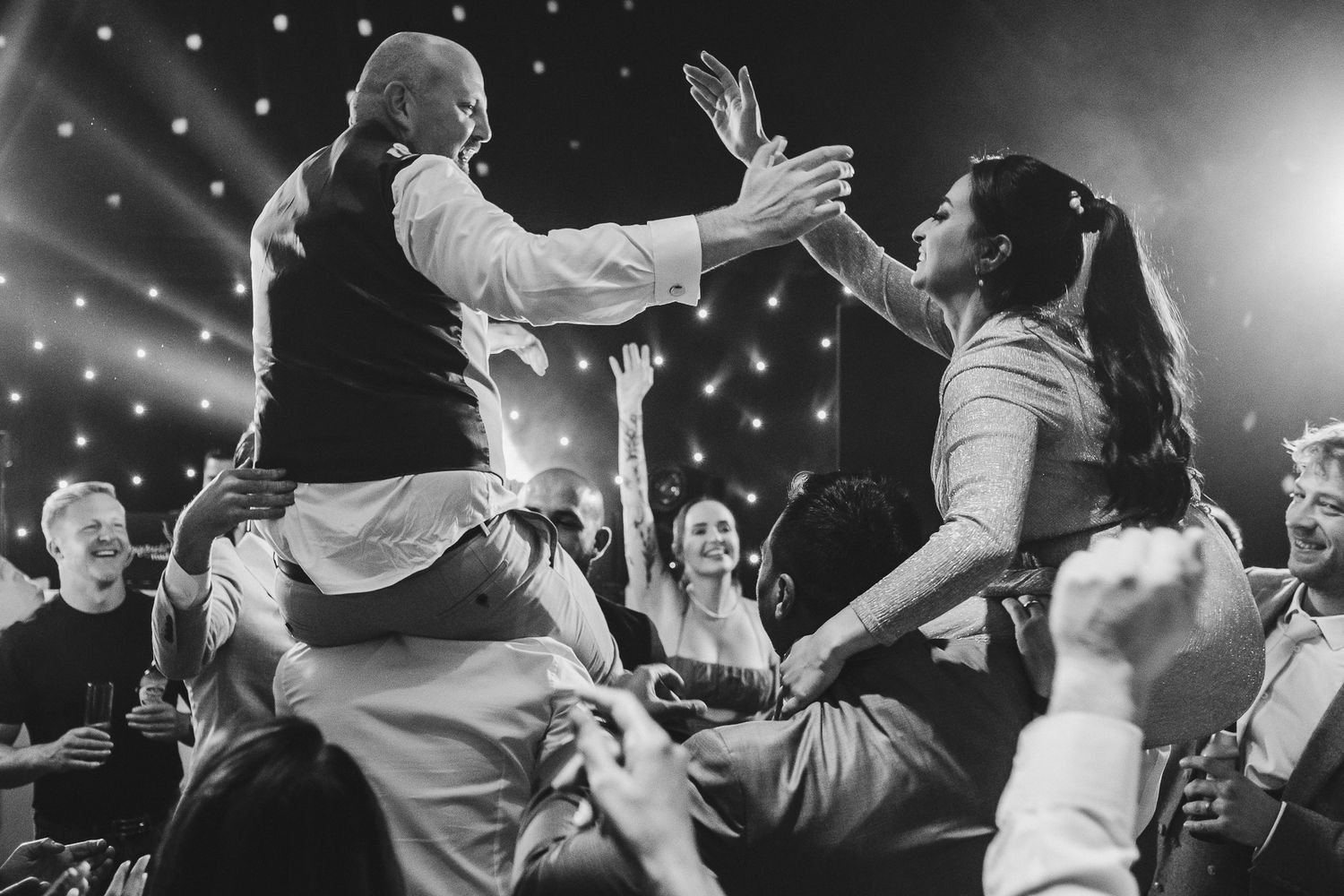 Two people in wedding attire dancing and celebrating energetically in front of a glittering background at a party reception.