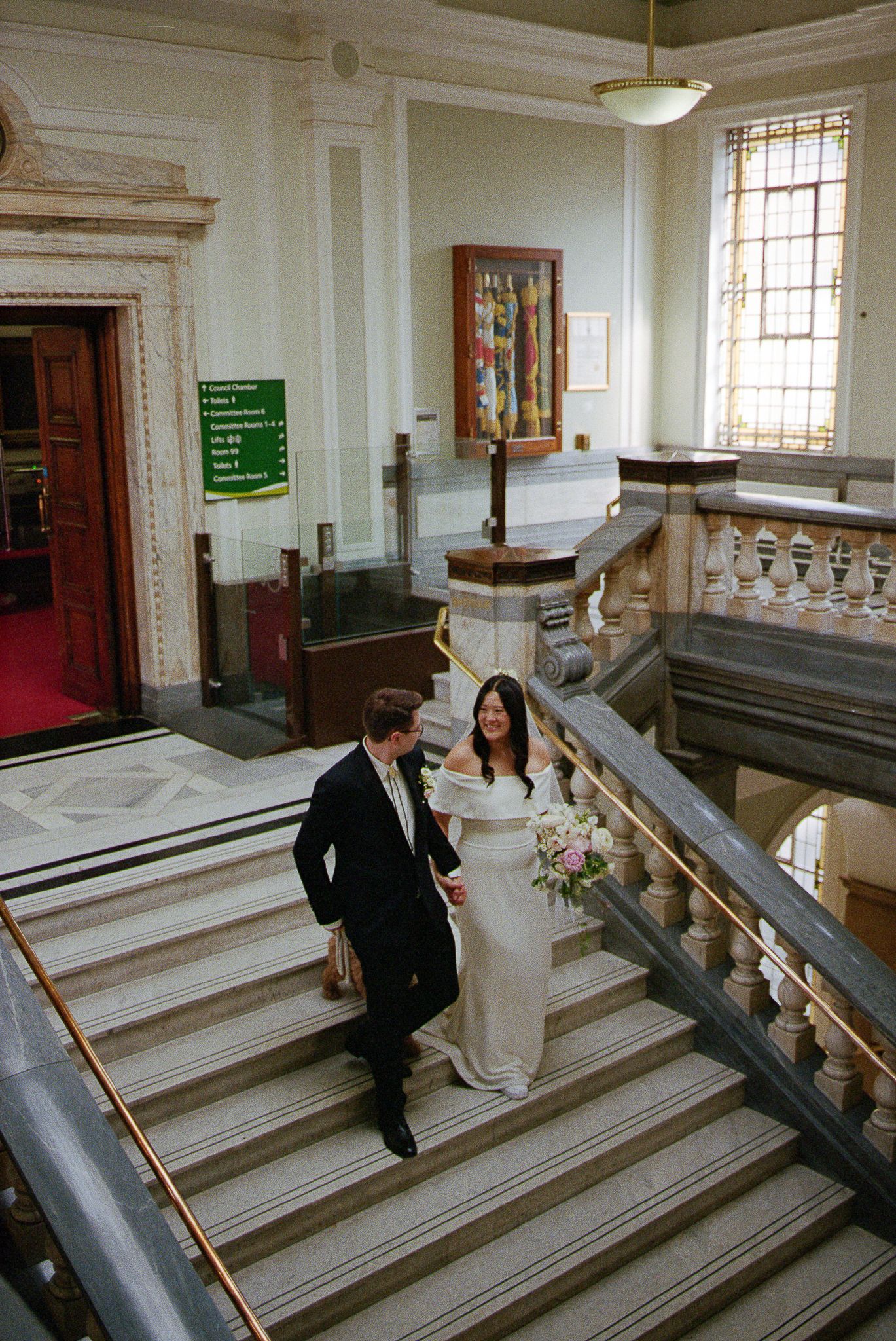 A couple in wedding attire walks down an elegant marble staircase inside a historic building with ornate architectural details.