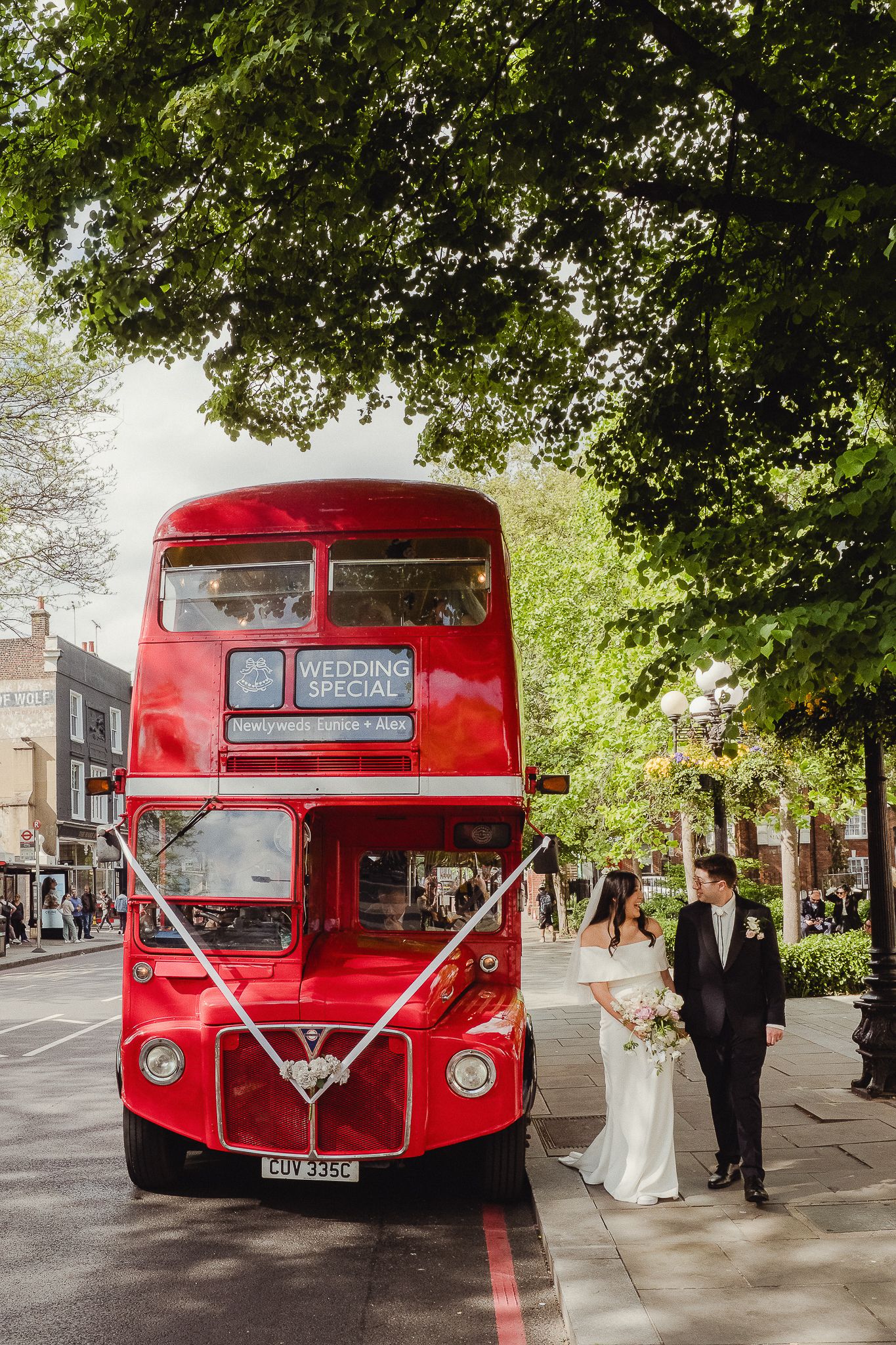 Classic red London double-decker bus decorated with white ribbons serves as wedding transport on a leafy city street.