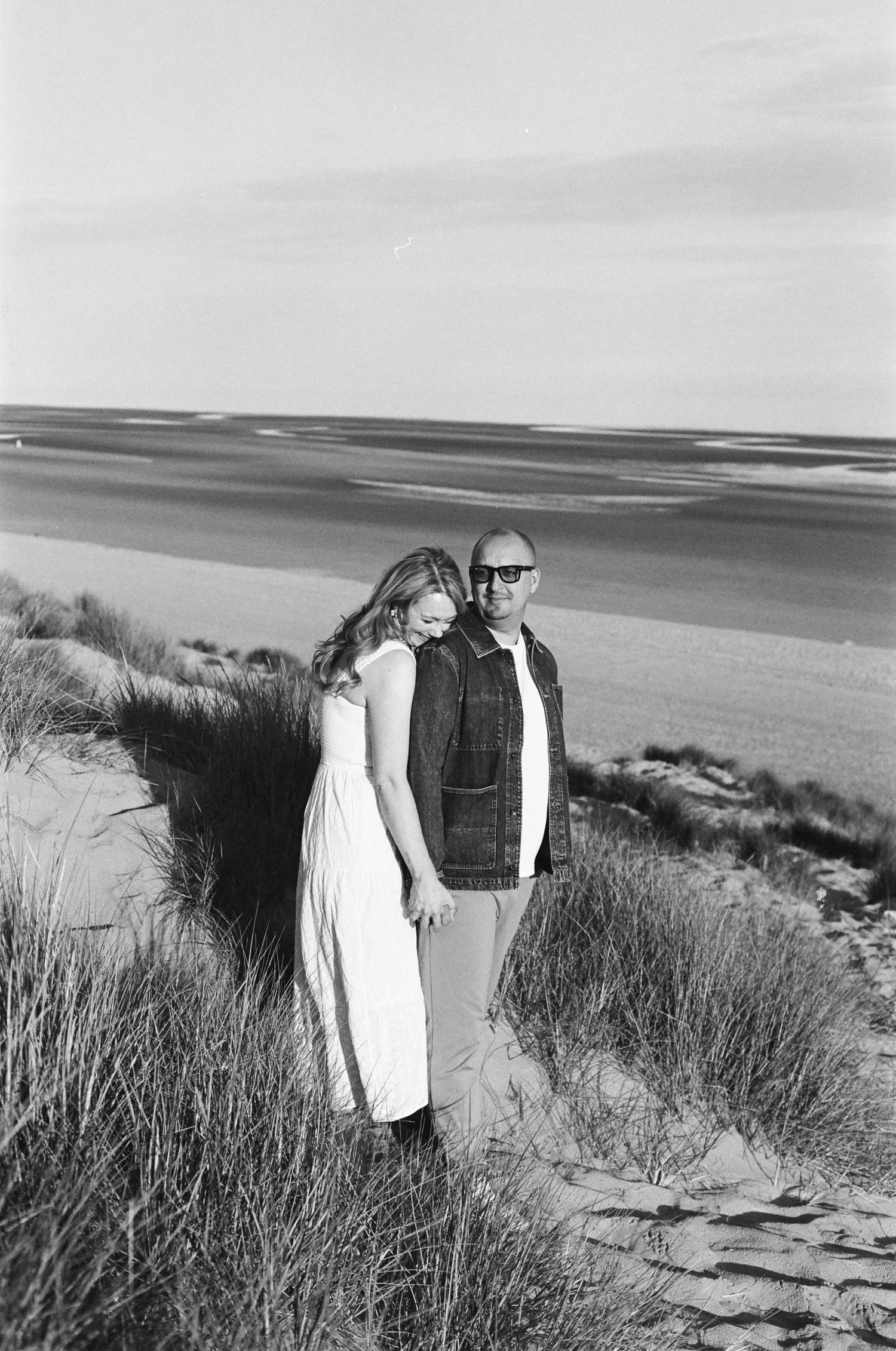 A black and white photograph shows two people standing together on grassy sand dunes overlooking the ocean.