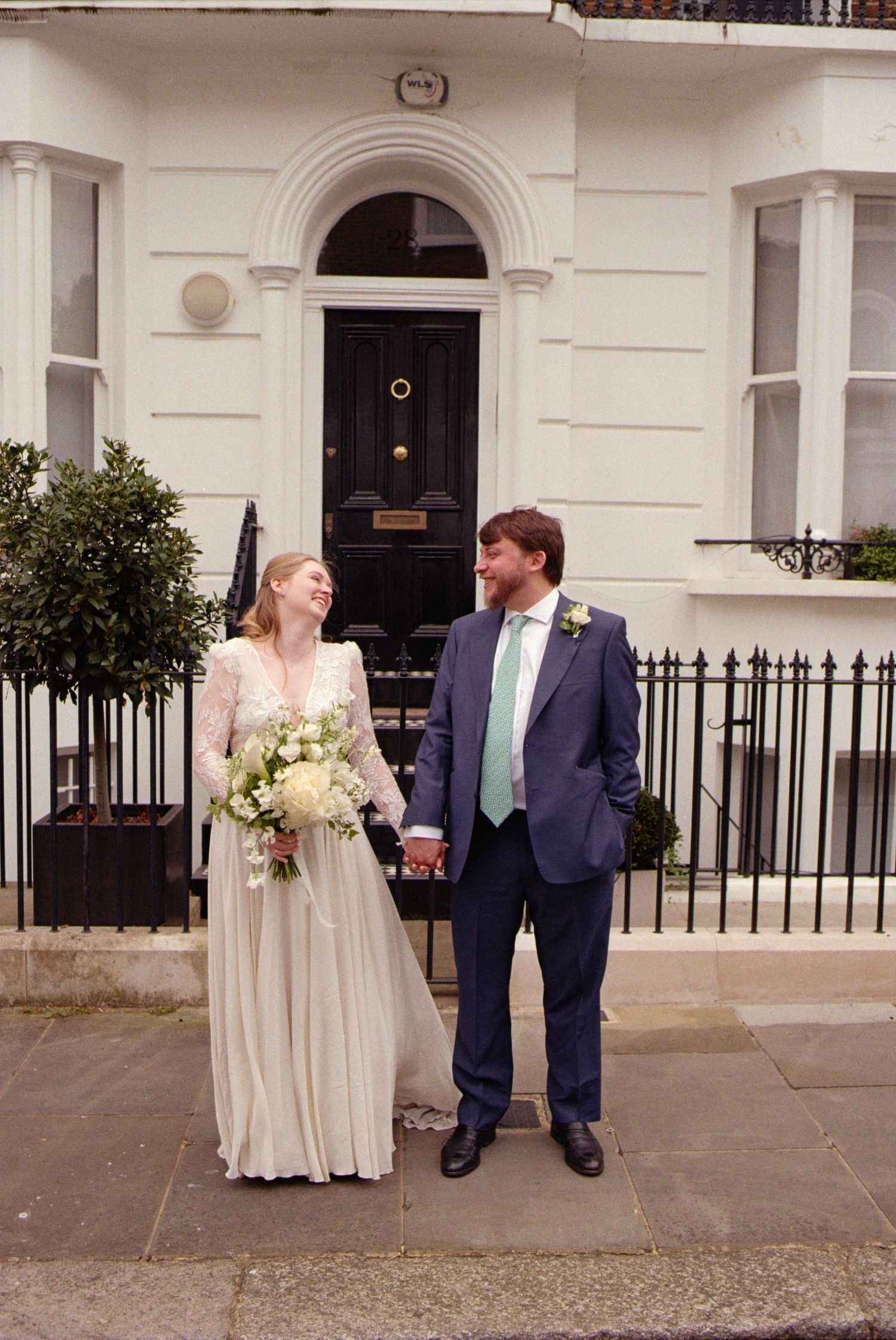 A couple stands smiling together on steps outside a classic white Georgian townhouse with black door and iron railings.