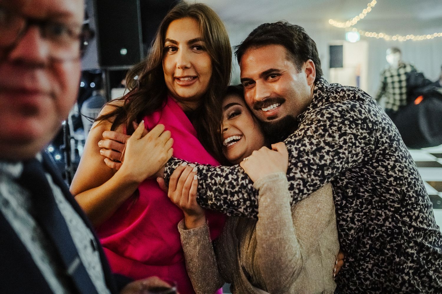 Three friends share a joyful moment together at an indoor gathering, with one wearing a bright pink dress and another in a patterned top.