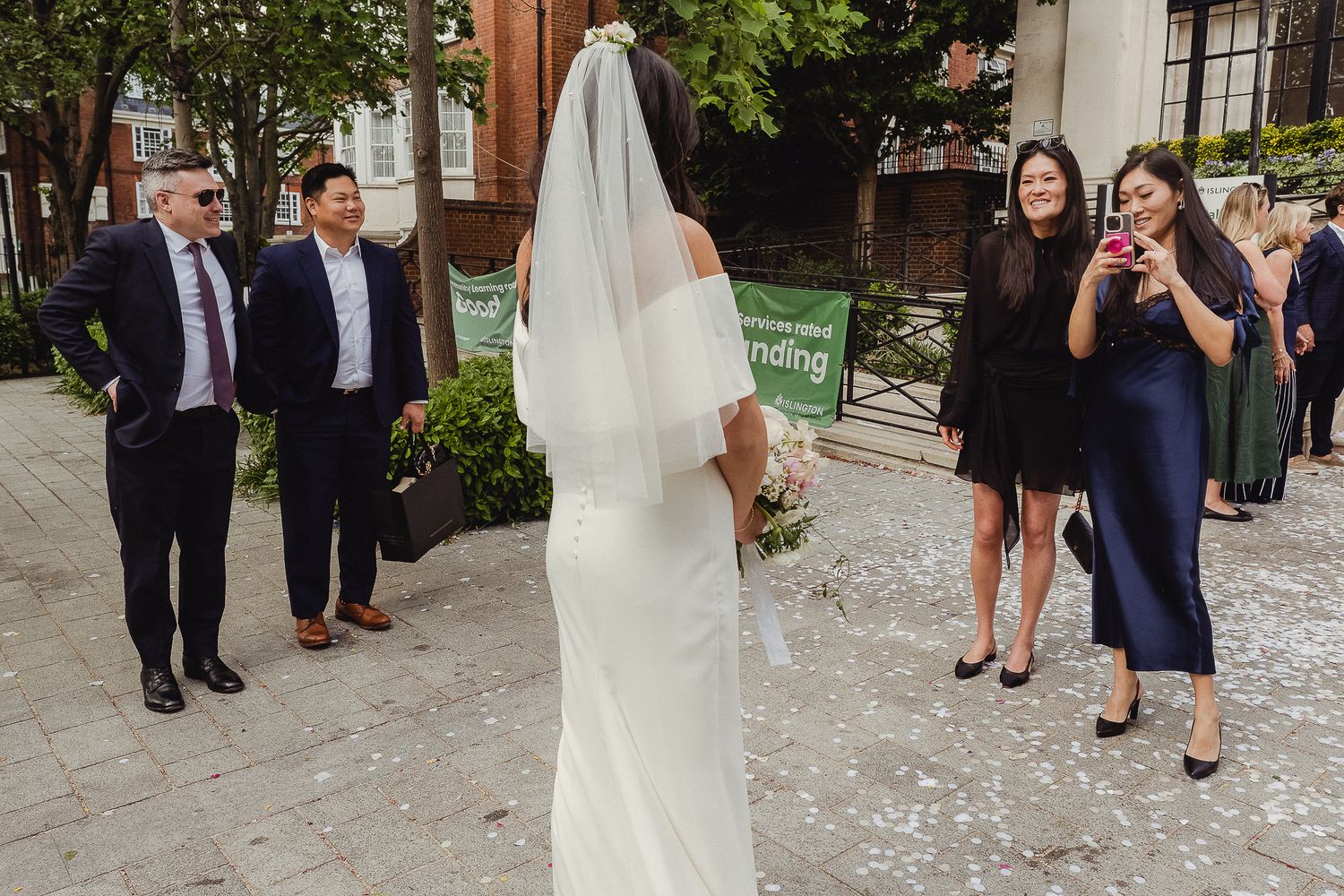 Wedding party members gather outside a brick building as a bride in a white gown and veil stands with her back to the camera.
