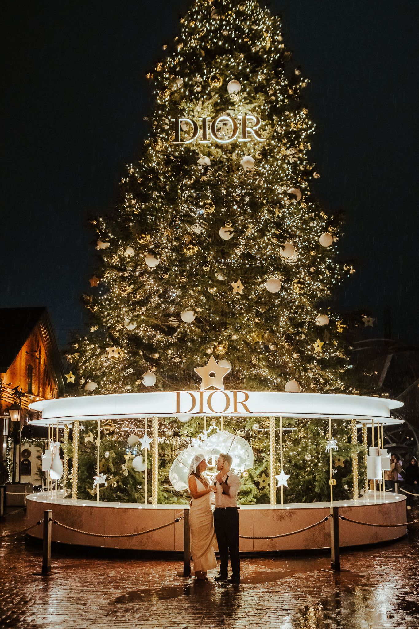 Couple stands beneath illuminated Dior Christmas tree display on rainy evening.
