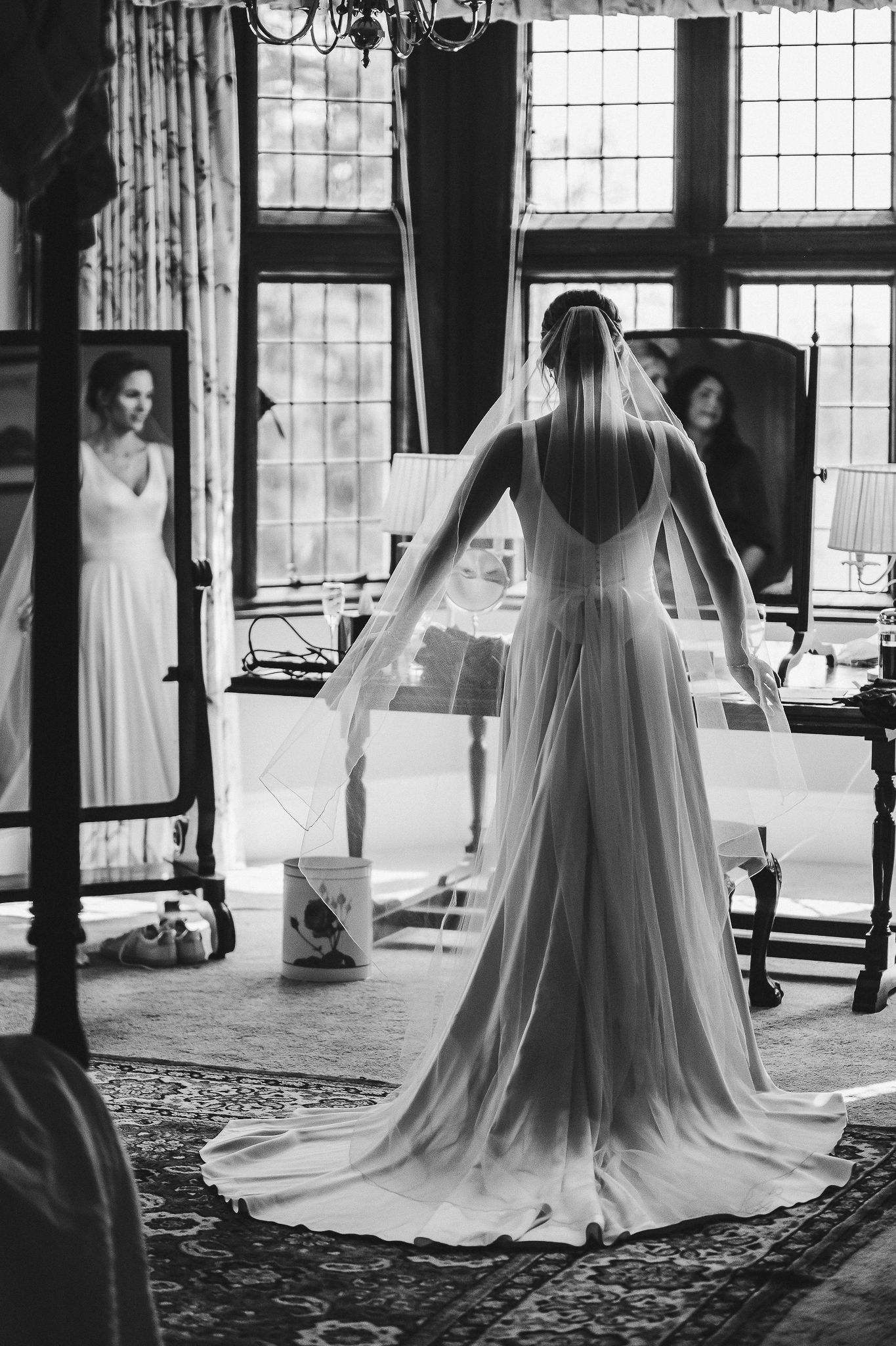 Bride in flowing gown viewed from behind looking at reflection in vintage mirror.
