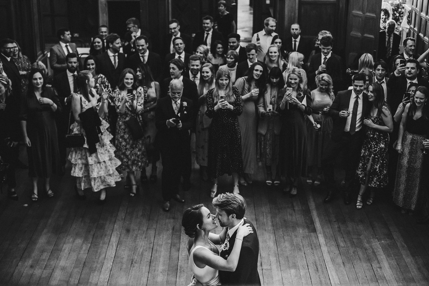 Wide shot of wedding reception crowd gathered in a grand hall with wooden floors.