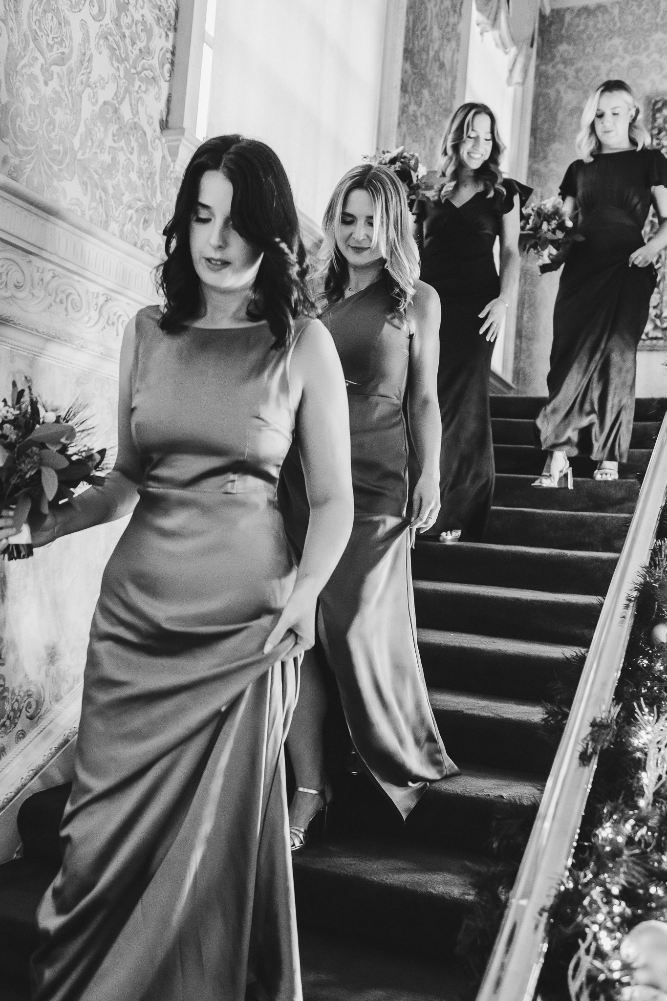 Bridesmaids in elegant satin dresses descending a vintage staircase.