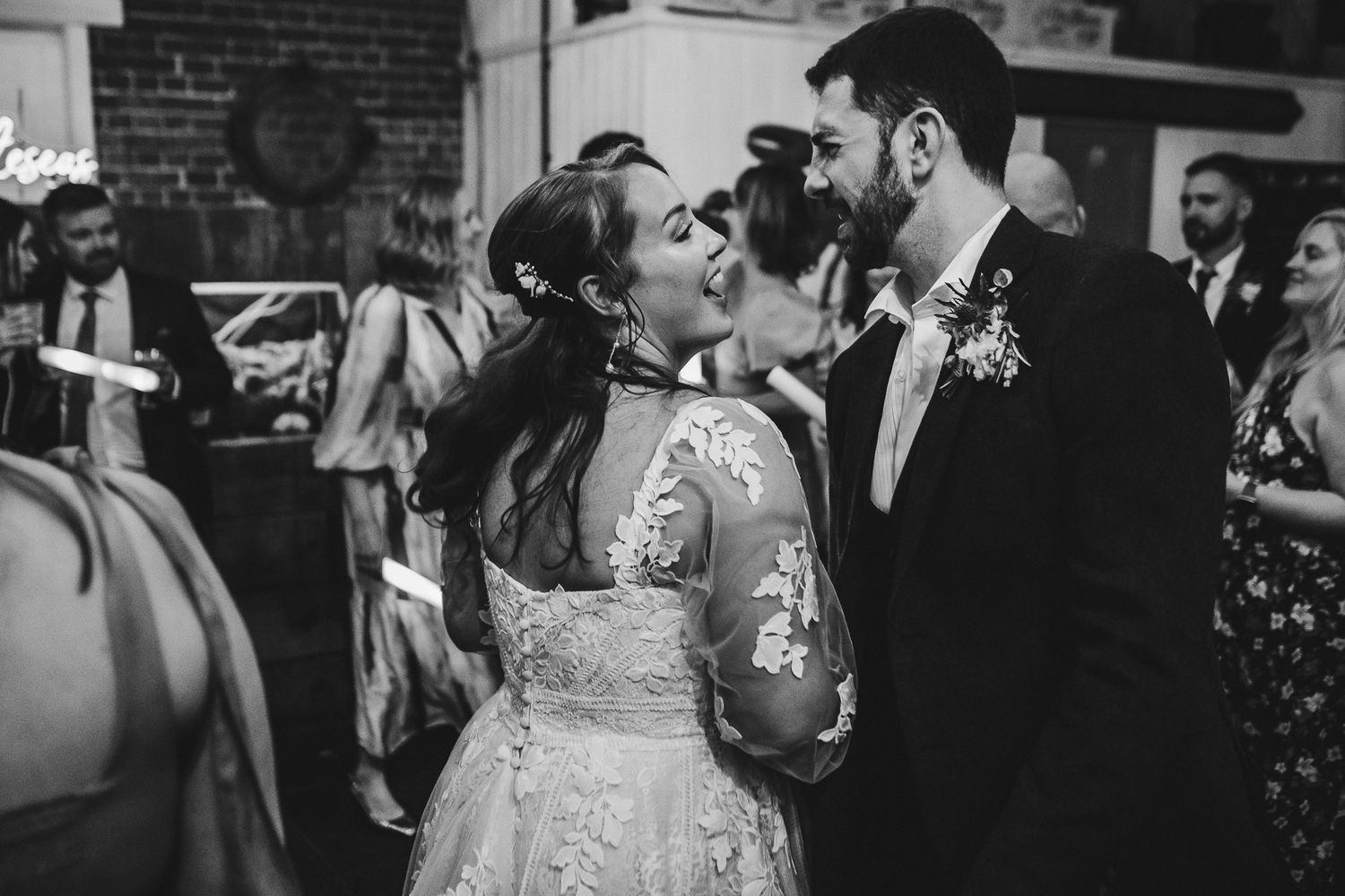 Wedding party guests dance and celebrate with glowing neon batons in a brick-walled venue.