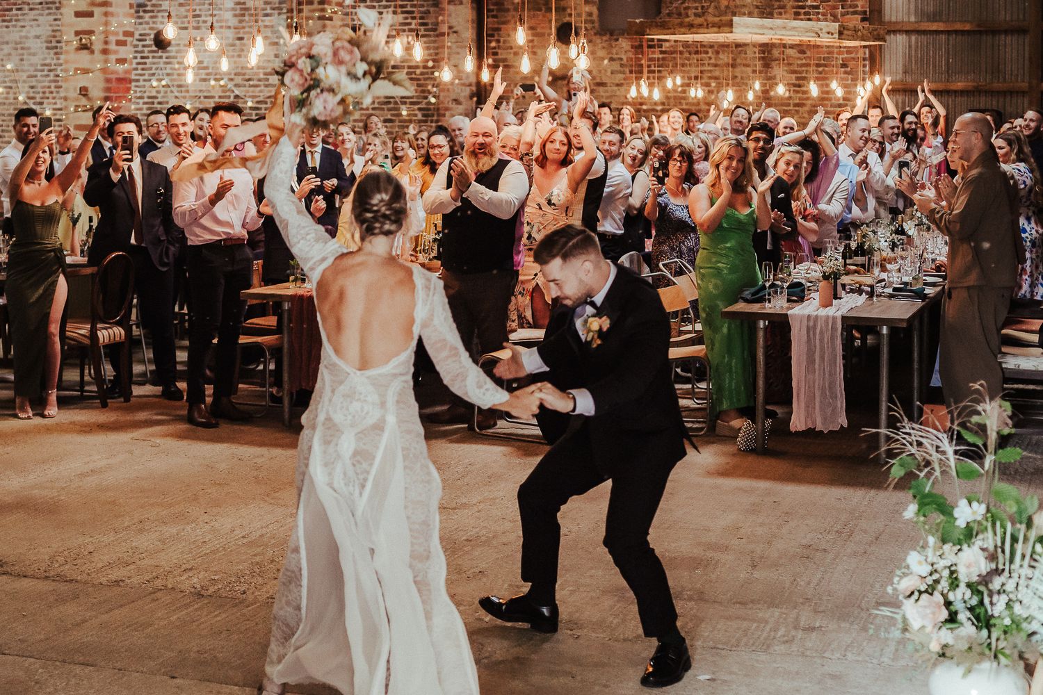 A couple performs an energetic dance move at their wedding reception while guests cheer and applaud in the background.