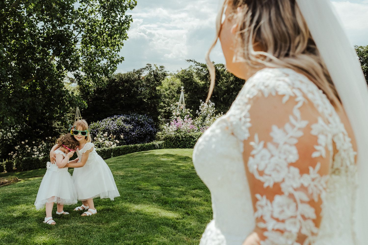 A flower girl in white dress twirls on a sunny lawn while a bride watches from the side.