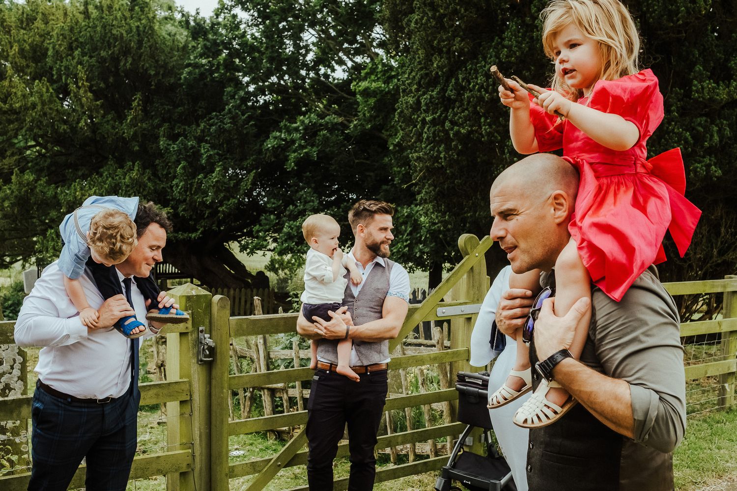 Wedding guests gather outdoors taking photos while a child in red sits on someone's shoulders.