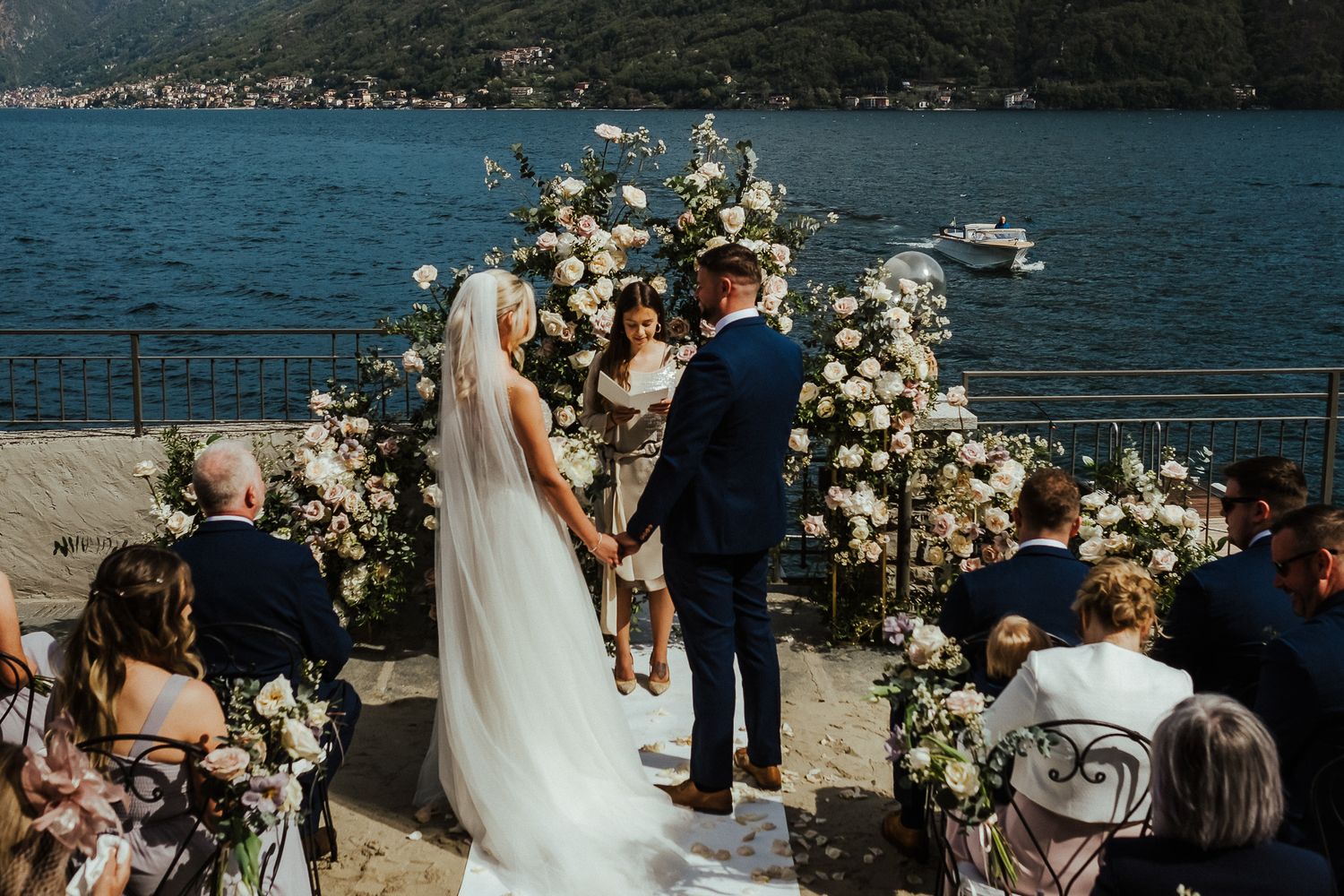 Couple exchanging vows at outdoor waterfront ceremony with mountain backdrop and floral arch.