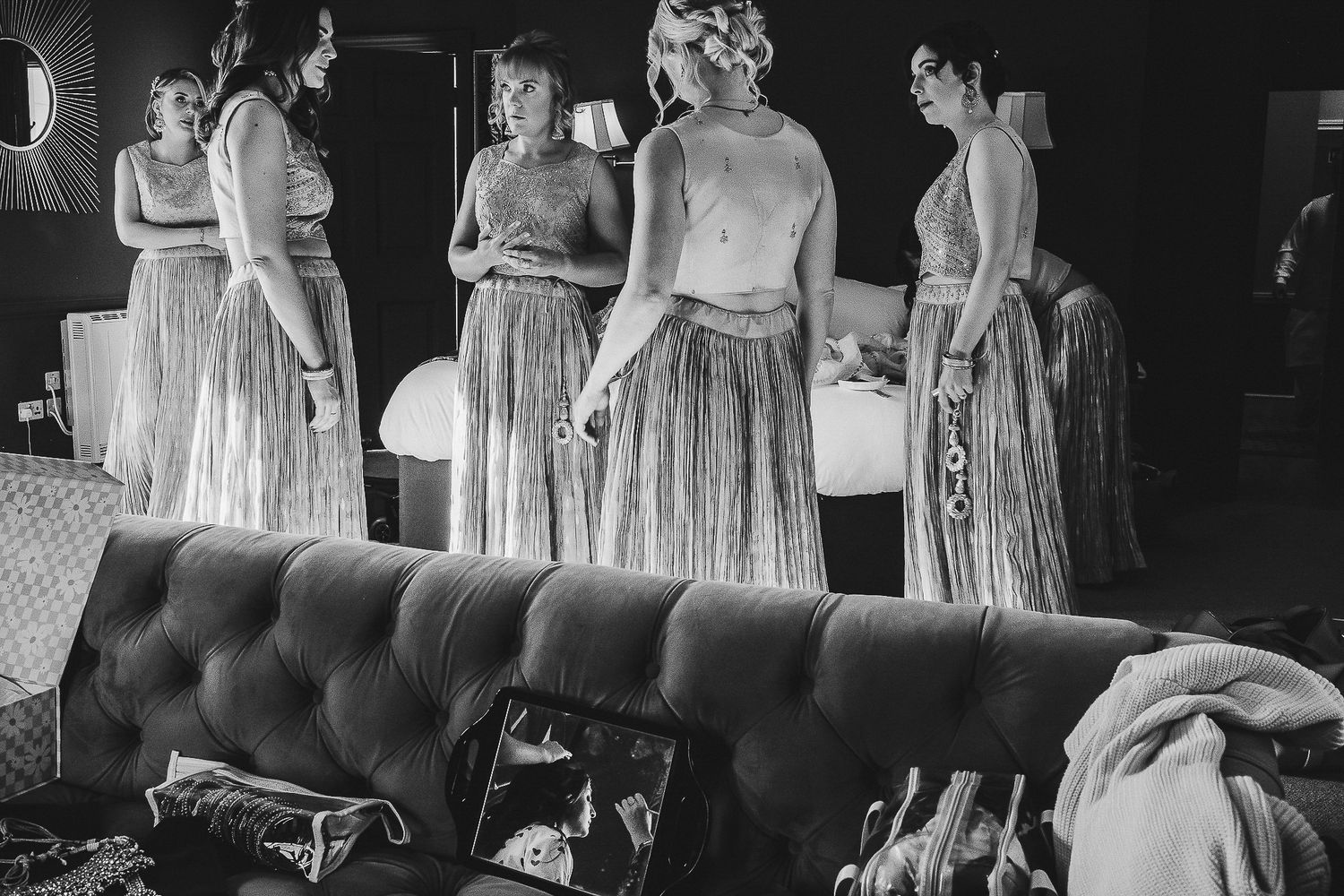 Bridesmaids in elegant flowy dresses prepare in a dressing room with a tufted couch in the foreground.