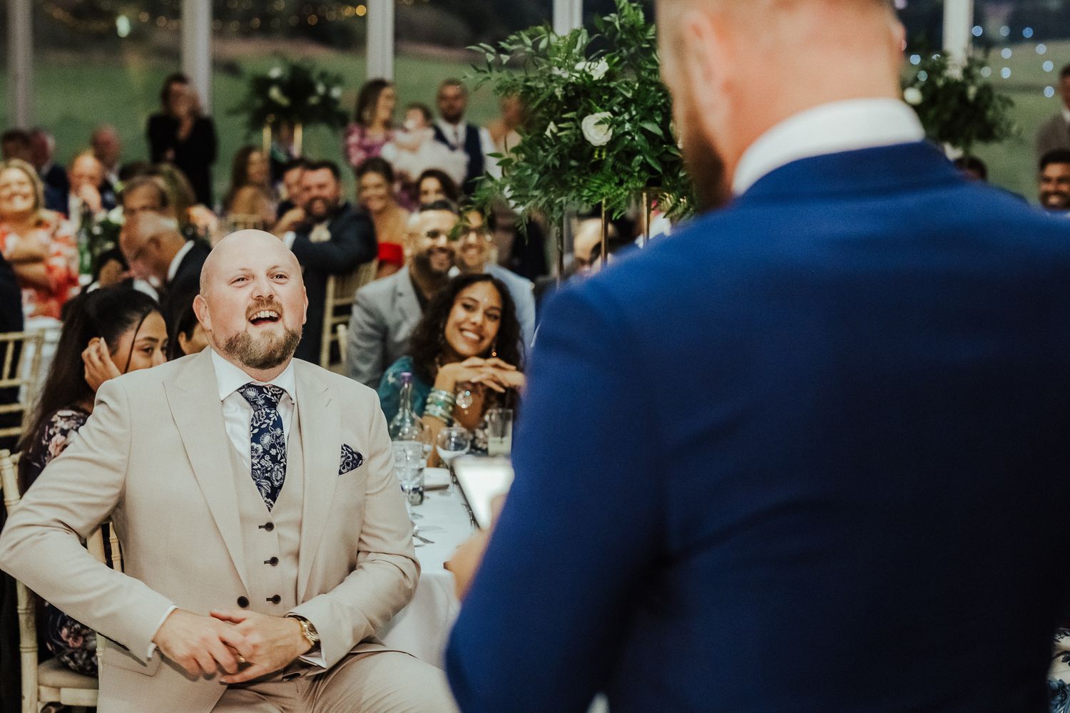 Guests at a wedding ceremony share joyful laughter during an indoor celebration with greenery decorations.