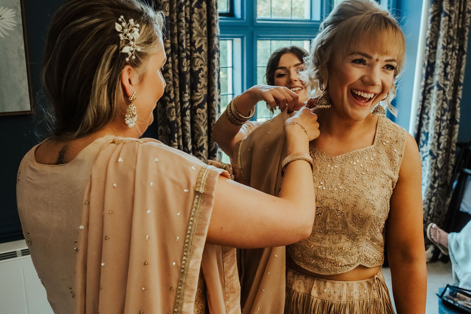 Bridesmaids help another bridesmaid get ready in a beige gown with decorative touches at an elegant indoor venue.