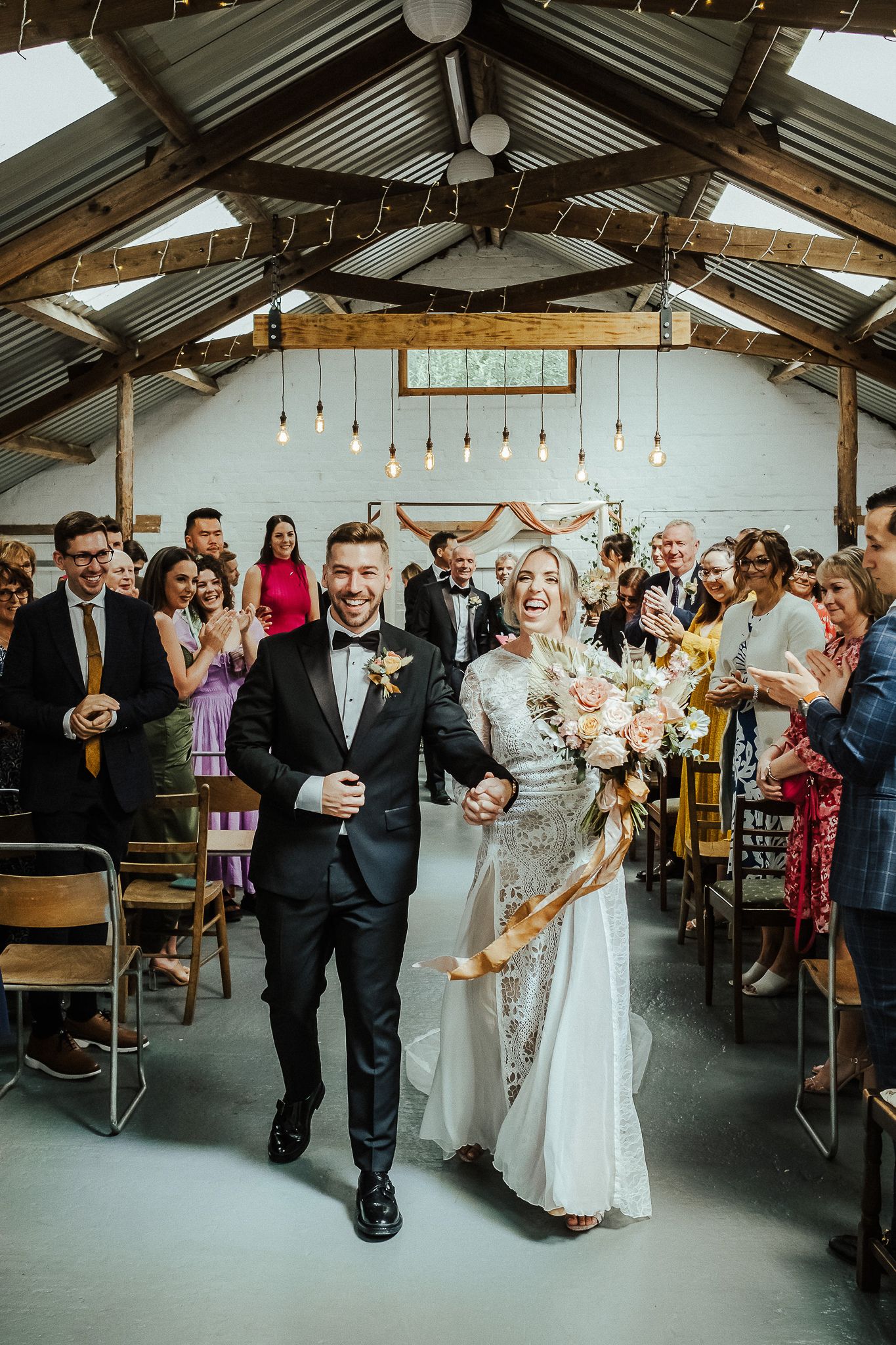 A joyful couple walks down the aisle of a rustic venue with exposed beams and pendant lights as guests celebrate.