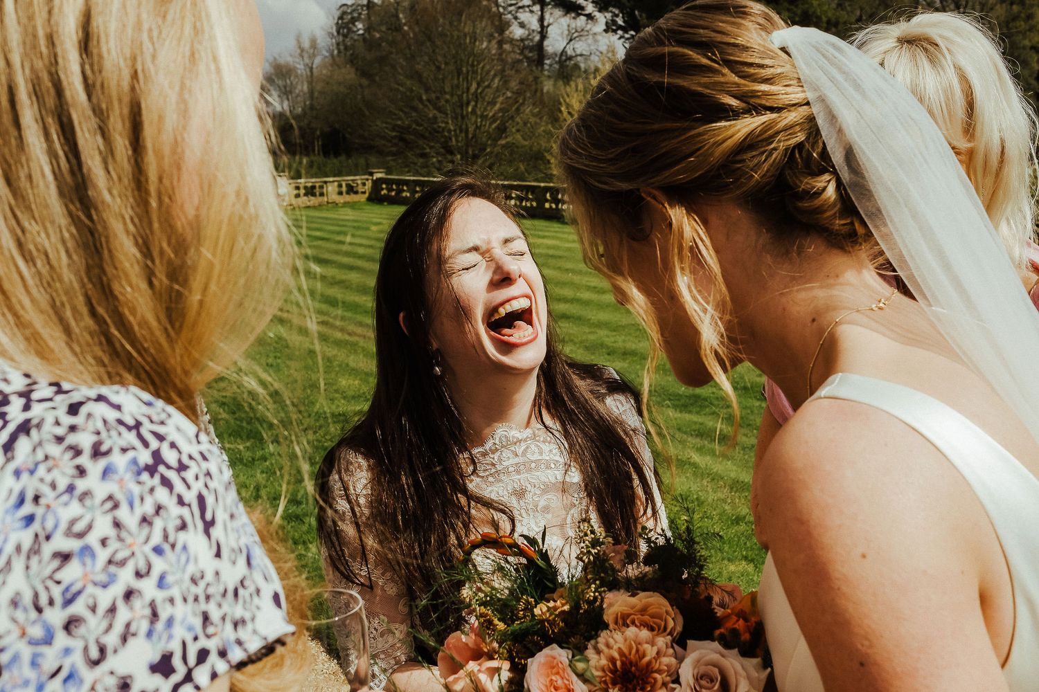 Three wedding attendees share a joyful laugh while holding bouquets outdoors.