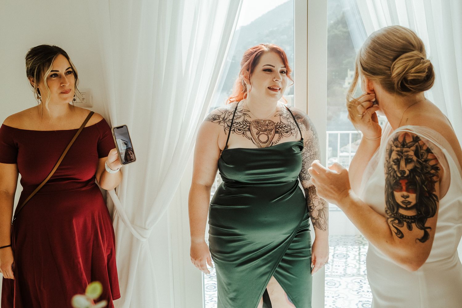 Three bridesmaids in elegant dresses share a candid moment by white curtains.