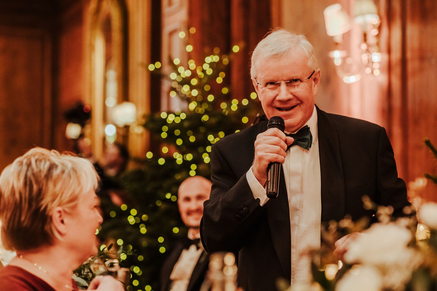 Speaker in bow tie addresses guests during formal holiday dinner event.