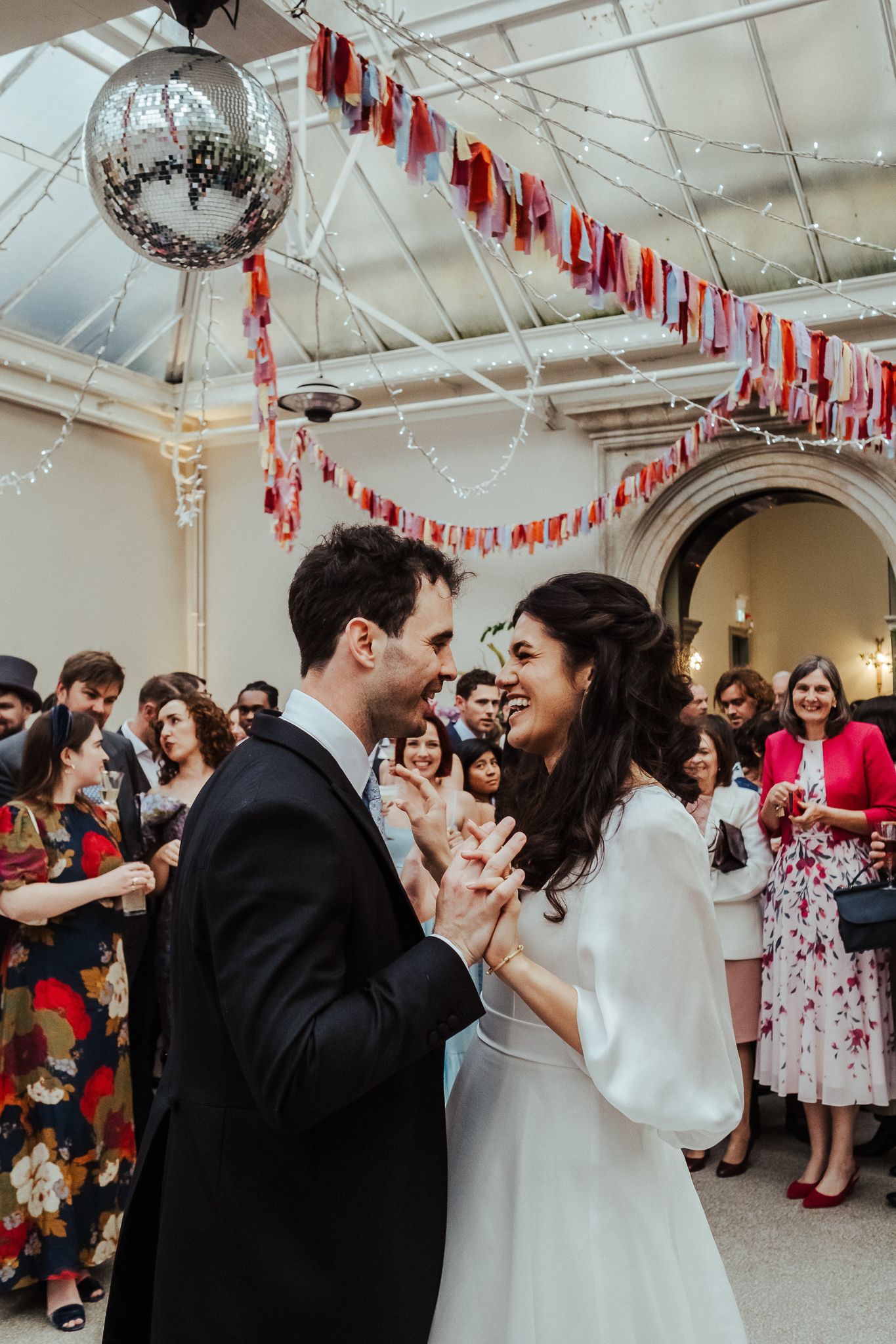 Newlyweds share their first dance under a disco ball and red streamers at an indoor reception.