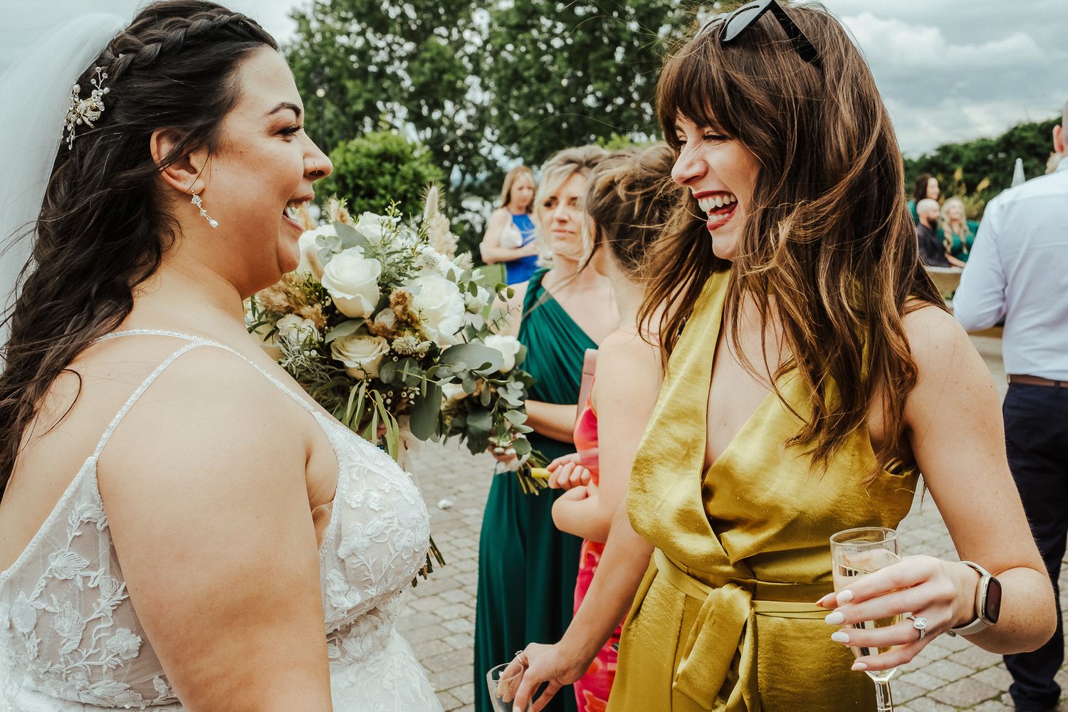 Two wedding guests share a candid laughing moment during an outdoor celebration.