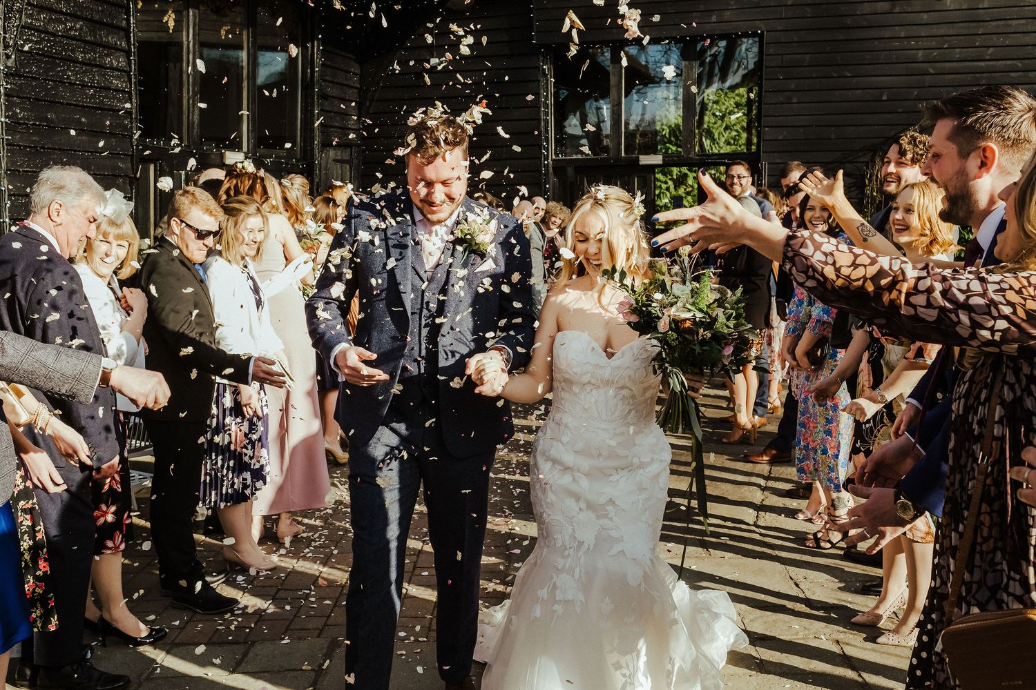 Wedding guests shower a couple with confetti as they exit a rustic venue.