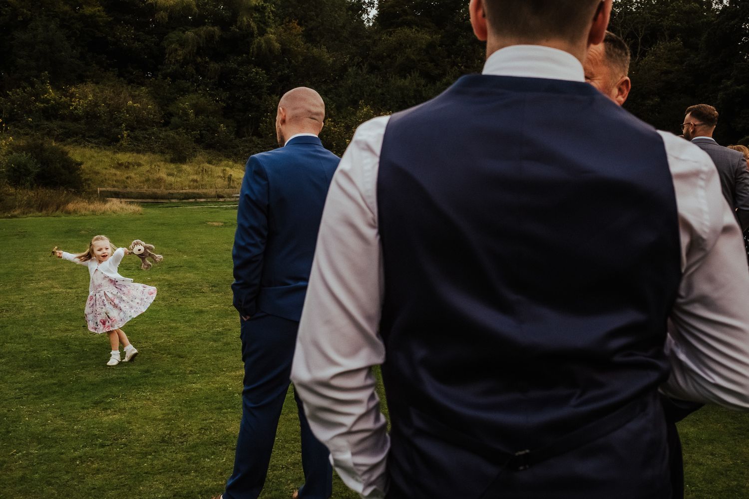 A bride wearing a white dress runs playfully through a green grassy field during a wedding celebration.